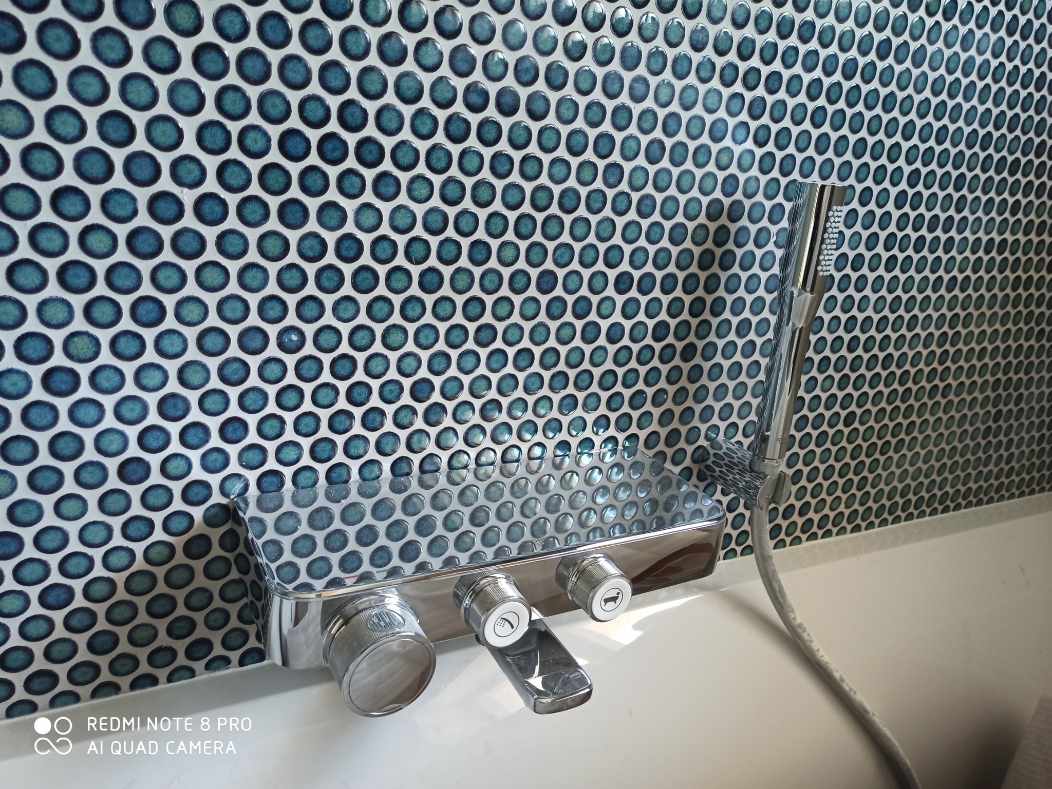 Bateria wannowa z chromowanym wykończeniem i słuchawką prysznicową na tle ściany wyłożonej mozaiką z drobnych, okrągłych płytek w odcieniach błękitu i bieli.