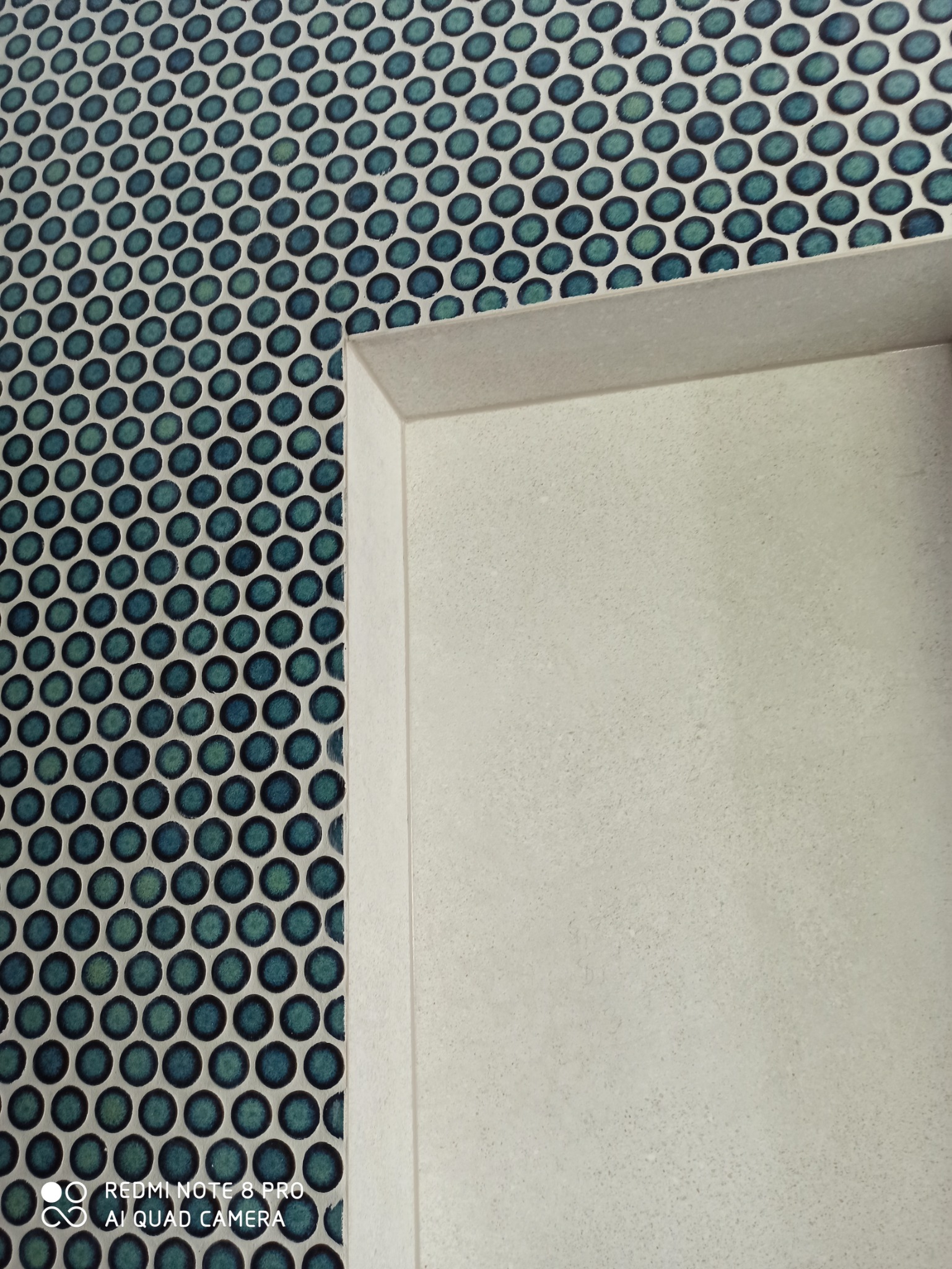 Fragment ściany wykończonej mozaiką z okrągłych, turkusowych płytek, sąsiadującej z wnęką wykończoną jasnym, matowym gresem.