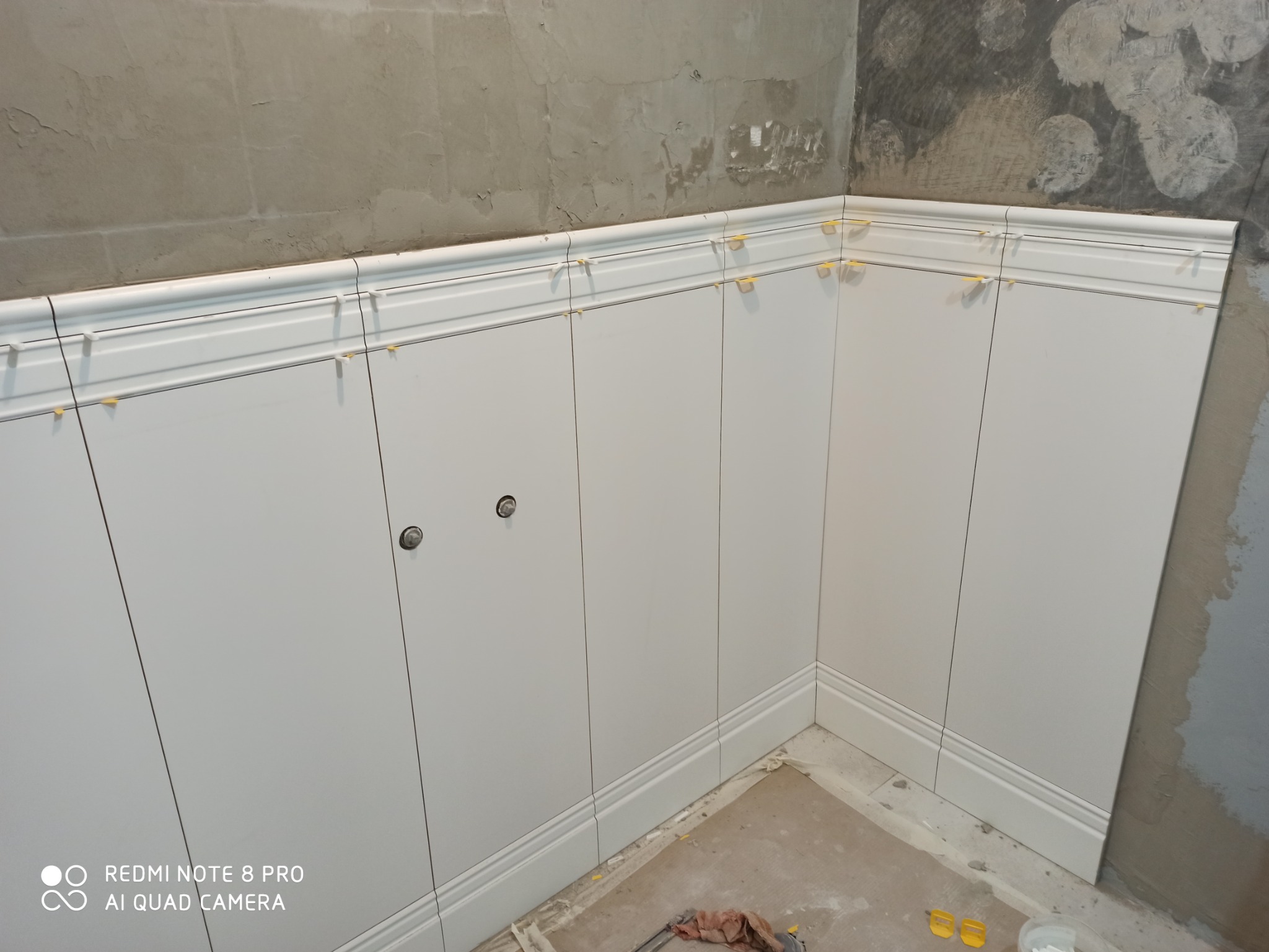 Białe panele ścienne z dekoracyjną listwą przypodłogową, zamontowane do połowy wysokości ściany, z widocznymi krzyżykami dystansowymi, na tle surowej, nieotynkowanej ściany.