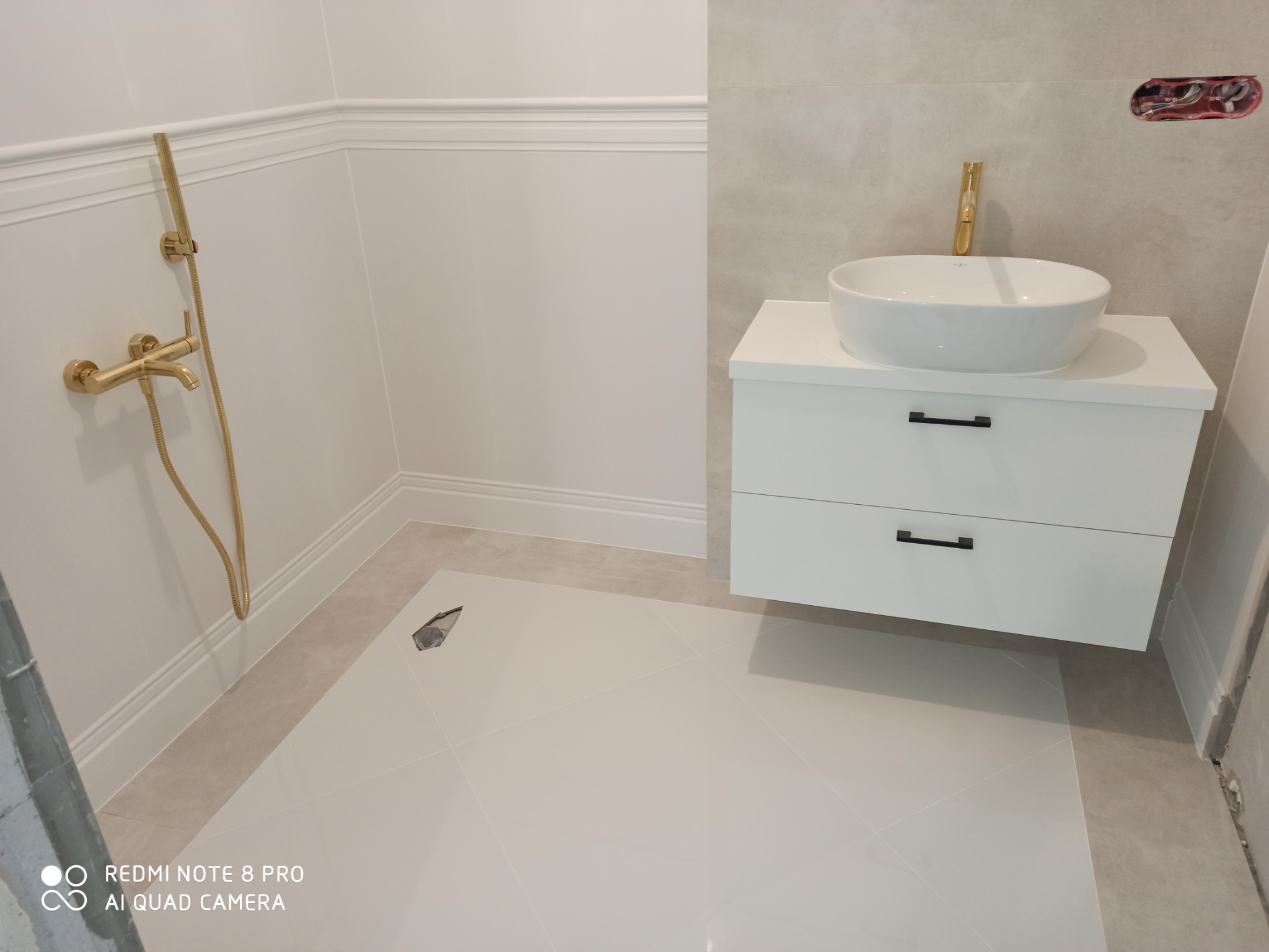 Nowoczesna łazienka w trakcie wykańczania, widoczna biała umywalka nablatowa z złotą baterią, złoty zestaw prysznicowy na ścianie, białe płytki podłogowe z widocznym otworem na odpływ, biała szafka...