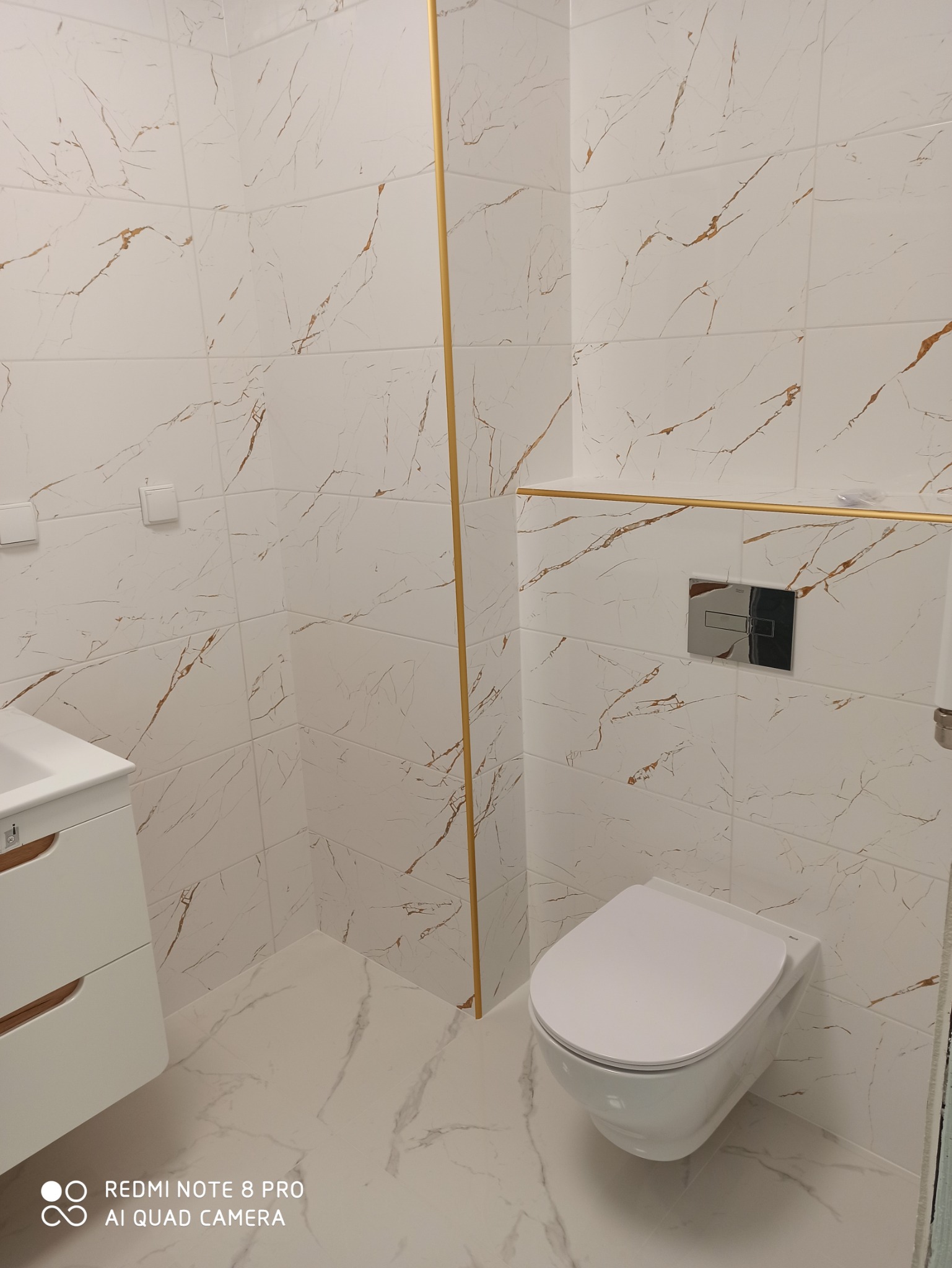 Wykończona łazienka z białymi płytkami imitującymi marmur z żyłkami w kolorze złota, wisząca toaleta z chromowanym przyciskiem spłukującym, biała szafka pod umywalkę z drewnianymi akcentami.