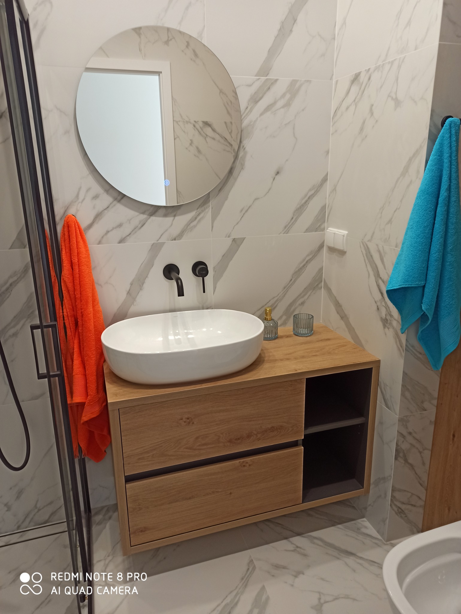 Nowoczesna łazienka z okrągłym lustrem z podświetleniem LED, białą umywalką nablatową na drewnianej szafce, czarną armaturą i marmurowymi płytkami ściennymi oraz pomarańczowym i turkusowym ręcznikiem.