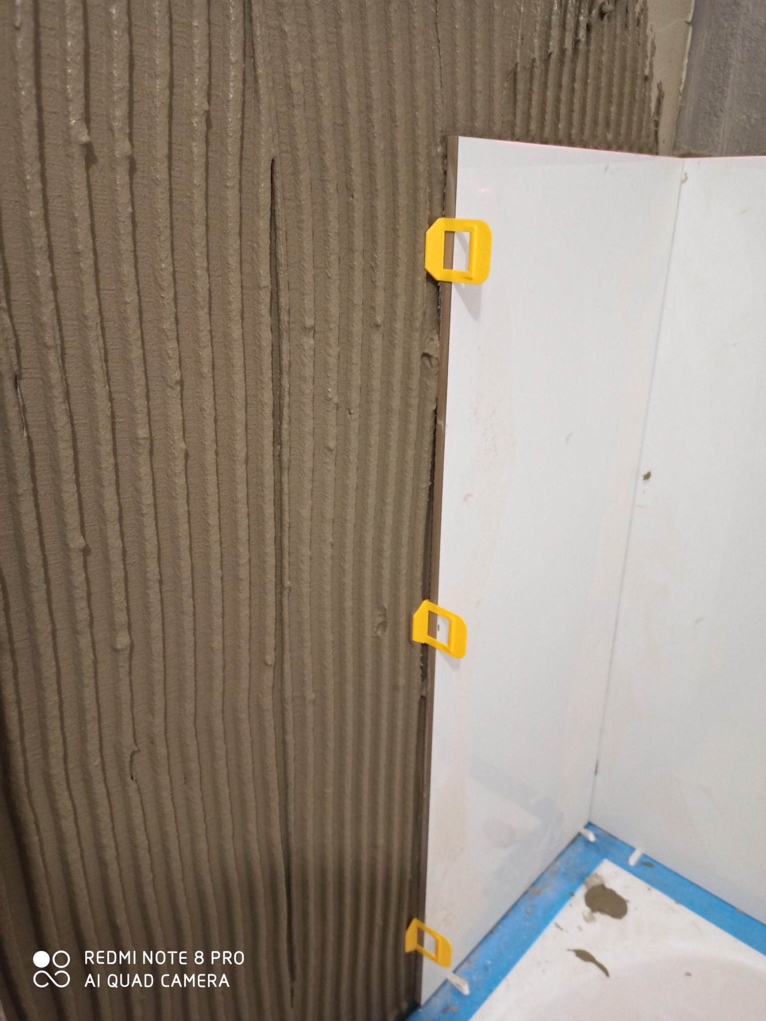 Świeżo ułożone, białe płytki na ścianie, z żółtymi krzyżykami dystansowymi, widoczna warstwa kleju z charakterystycznymi rowkami, zabezpieczona wanna.