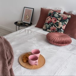 EcoProperty sp. z o.o. - Jasne łóżko z dekoracyjnymi poduszkami w odcieniach różu i beżu, drewniana taca z dwoma różowymi kubkami i rogalikiem, obok stolik z dekoracją, całość w jasnym, minimalistycznym wnętrzu.
