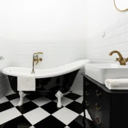 Elegancka łazienka z wolnostojącą wanną na lwich łapach, biała umywalka na czarnej komodzie z mosiężnymi uchwytami, czarno-biała szachownica na podłodze i białe kafelki na ścianach.
