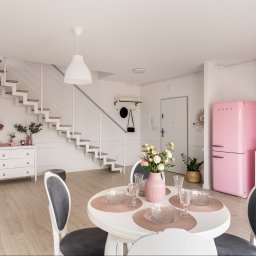 EcoProperty sp. z o.o. - Jasne, przestronne wnętrze z białymi ścianami, drewnianą podłogą, okrągłym stołem z nakryciem, różowym wazonem z kwiatami, białymi krzesłami z szarymi siedziskami, białymi schodami i różową lodówką...