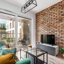 EcoProperty sp. z o.o. - Salon z ceglaną ścianą, szklanym stolikiem kawowym i wyjściem na balkon, widok na nowoczesny budynek z zewnątrz.