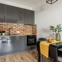 EcoProperty sp. z o.o. - Szara kuchnia z ceglaną ścianą, czarnym zlewem i baterią, drewnianym blatem, z roślinami w doniczkach, oraz czarny stół z żółtym bieżnikiem, na którym leżą rogaliki i woda z cytryną.