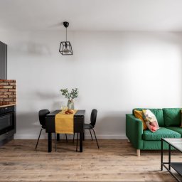 EcoProperty sp. z o.o. - Jasne wnętrze mieszkania: szare szafki kuchenne z ceglanym backsplash'em, czarny stół z żółtym bieżnikiem, zielona sofa z kolorowymi poduszkami, drewnopodobna podłoga, czarne lampy sufitowe.