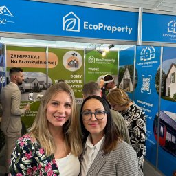 EcoProperty sp. z o.o. - Dwie uśmiechnięte kobiety na tle stoiska targowego EcoProperty z wizualizacjami domów i informacjami o metrażu, z widocznym logo firmy i QR kodem.