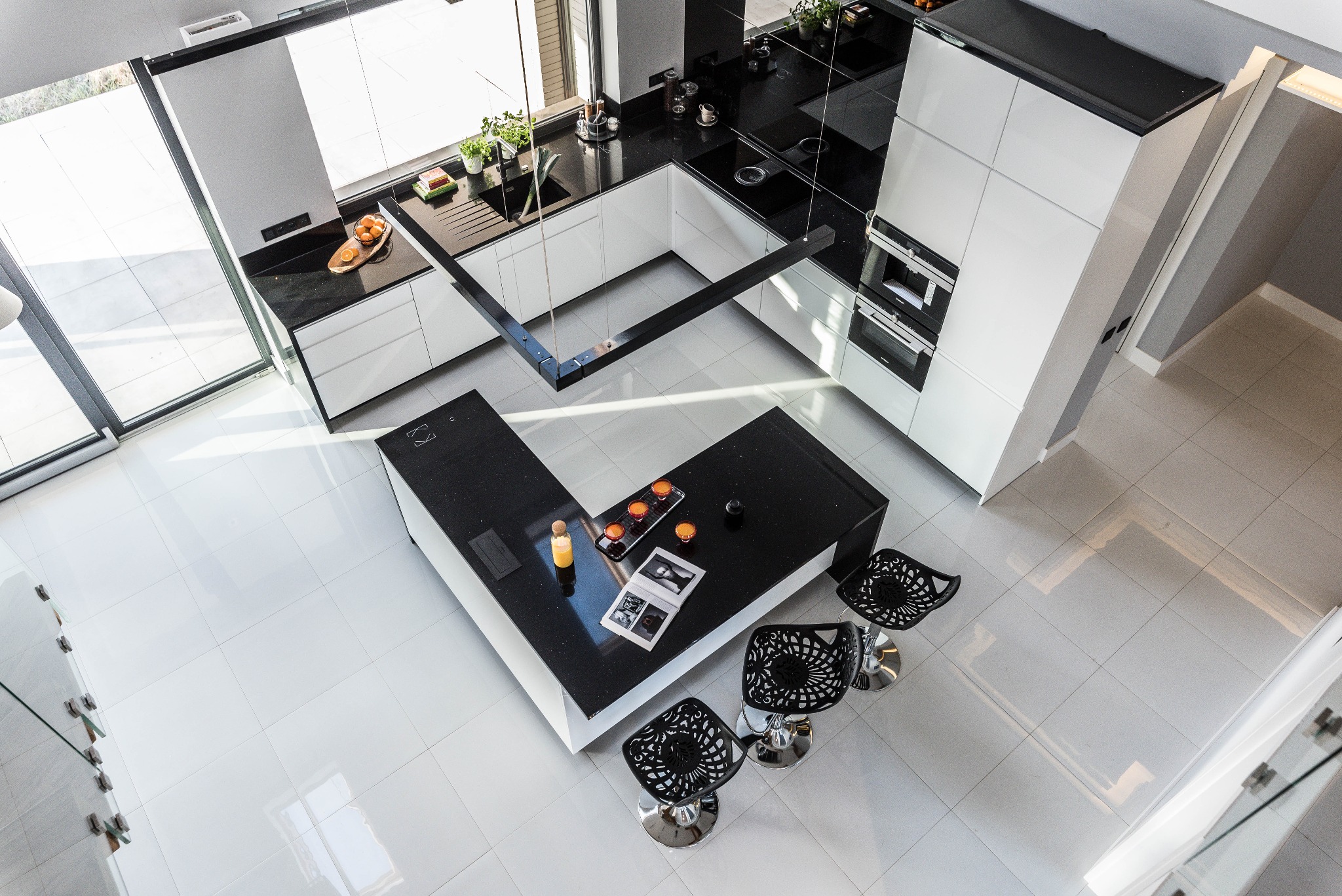 Nowoczesna kuchnia z wyspą, czarne blaty i białe szafki, widok z góry na minimalistyczne krzesła barowe i akcesoria dekoracyjne na blacie.