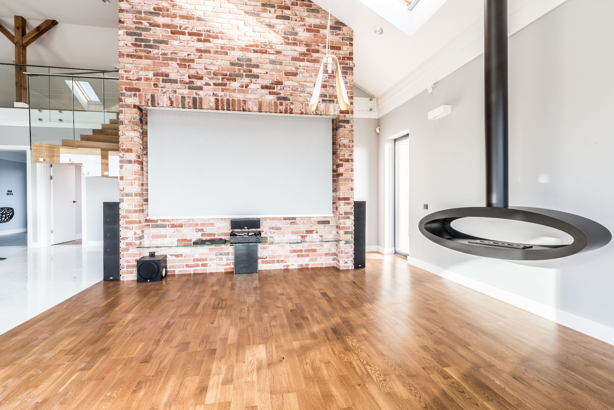Przestronny salon z parkietem, ceglaną ścianą z ekranem projekcyjnym, nowoczesnym kominkiem podwieszanym i schodami na antresolę z drewnianymi stopniami.