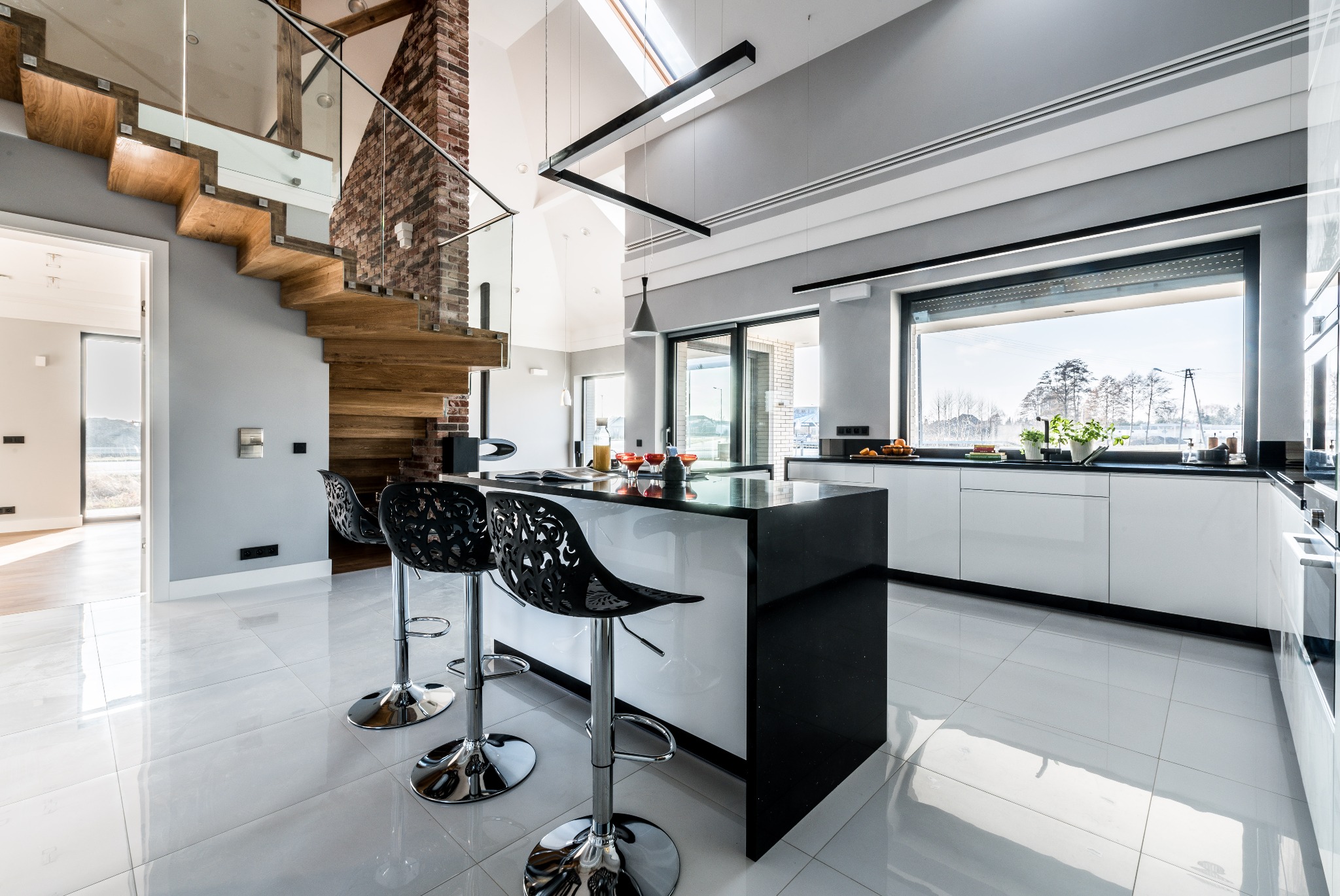 Nowoczesna, jasna kuchnia z wyspą i krzesłami barowymi o ażurowych oparciach, drewniane schody z elementami cegły i szklaną balustradą, duże okno z widokiem na zimowy krajobraz.
