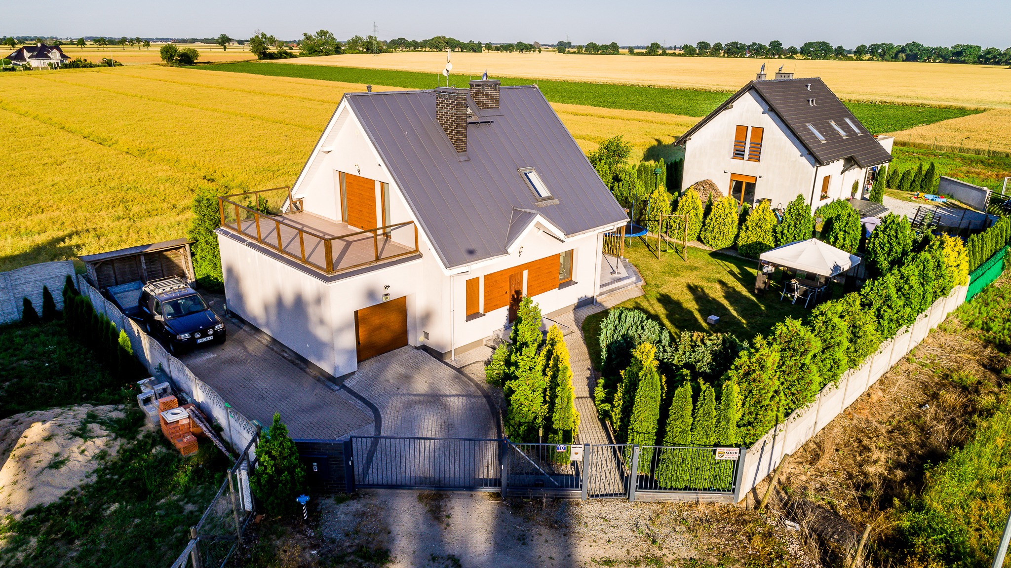Dwa nowoczesne domy jednorodzinne z szarymi dachami i drewnianymi elementami, widok z góry, otoczone zielonymi tujami i polami uprawnymi.