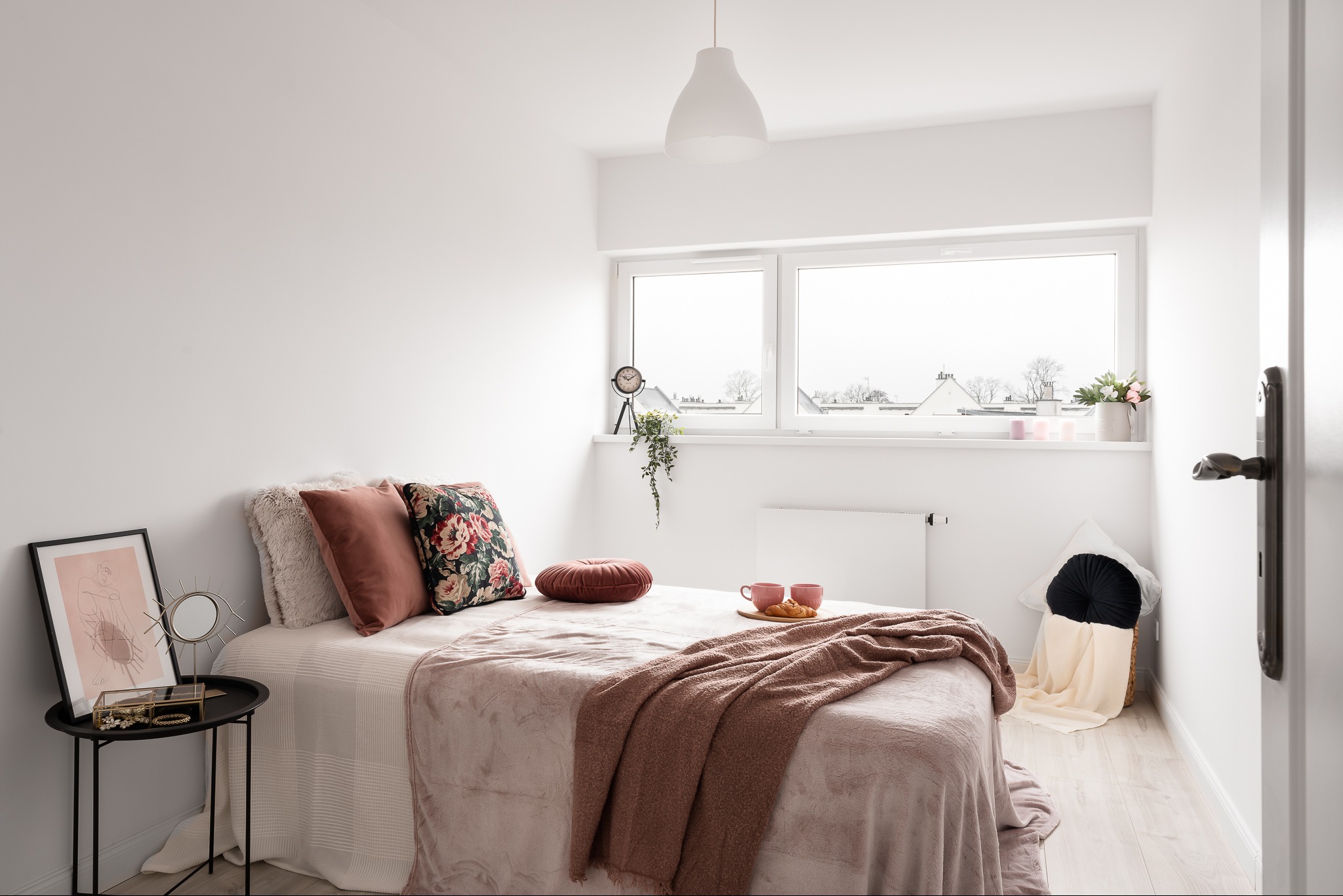 Jasna sypialnia z łóżkiem, stolikiem nocnym z dekoracjami, widokiem z okna i elementami home stagingu, w tym poduszki, pled i śniadanie na łóżku.
