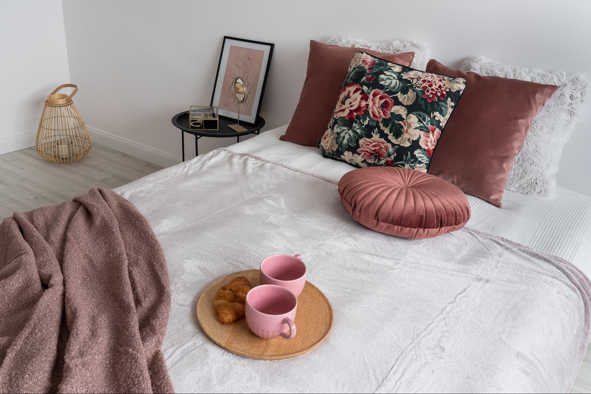 Jasne łóżko z dekoracyjnymi poduszkami w odcieniach różu i beżu, drewniana taca z dwoma różowymi kubkami i rogalikiem, obok stolik z dekoracją, całość w jasnym, minimalistycznym wnętrzu.
