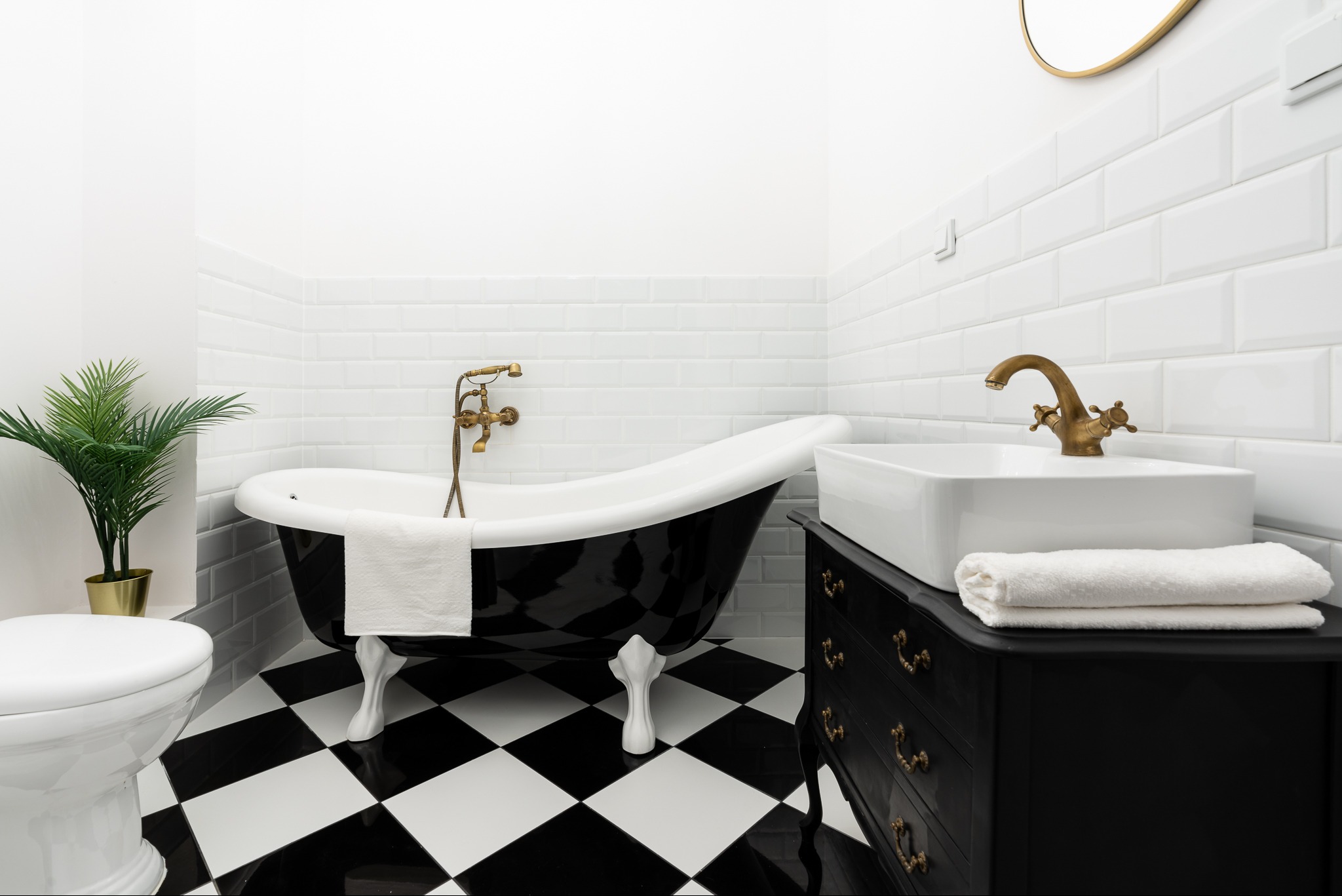 Elegancka łazienka z wolnostojącą wanną na lwich łapach, biała umywalka na czarnej komodzie z mosiężnymi uchwytami, czarno-biała szachownica na podłodze i białe kafelki na ścianach.