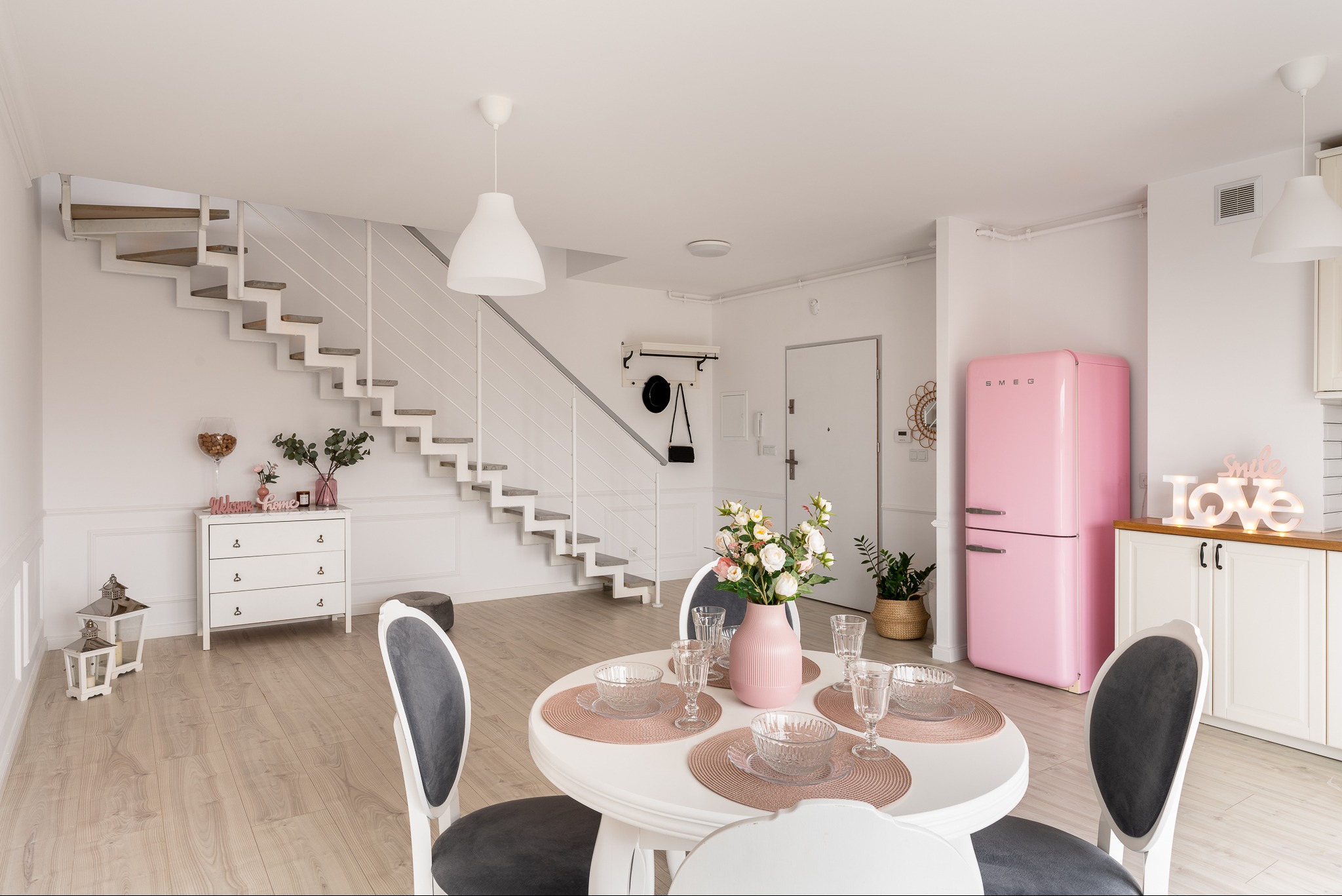 Jasne, przestronne wnętrze z białymi ścianami, drewnianą podłogą, okrągłym stołem z nakryciem, różowym wazonem z kwiatami, białymi krzesłami z szarymi siedziskami, białymi schodami i różową lodówką...