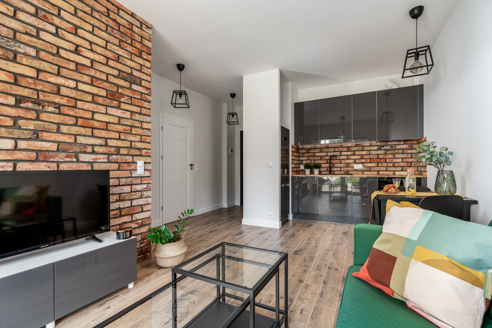 Nowoczesne wnętrze mieszkania z ceglaną ścianą, szarą zabudową kuchenną i elementami dekoracyjnymi, takimi jak roślina w koszyku i geometryczna poduszka na zielonej sofie.