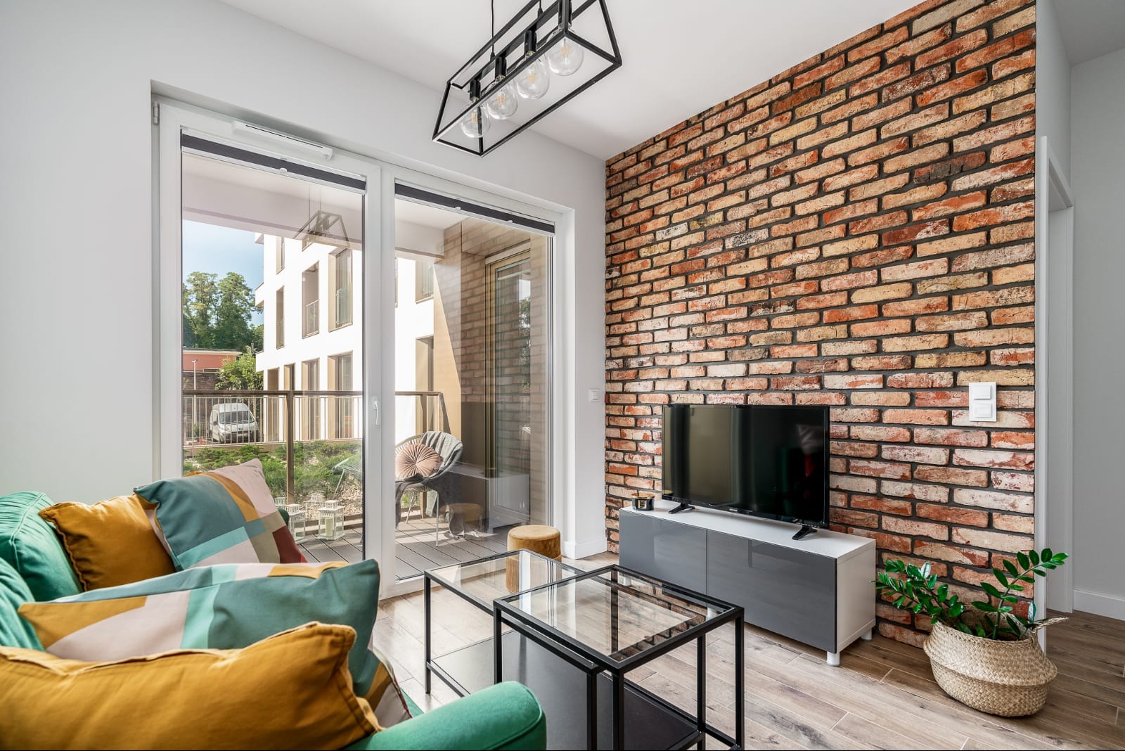 Salon z ceglaną ścianą, szklanym stolikiem kawowym i wyjściem na balkon, widok na nowoczesny budynek z zewnątrz.
