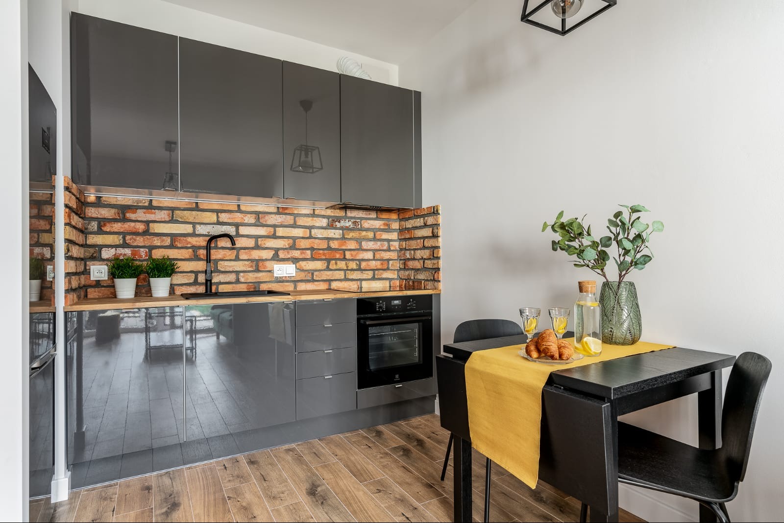 Szara kuchnia z ceglaną ścianą, czarnym zlewem i baterią, drewnianym blatem, z roślinami w doniczkach, oraz czarny stół z żółtym bieżnikiem, na którym leżą rogaliki i woda z cytryną.