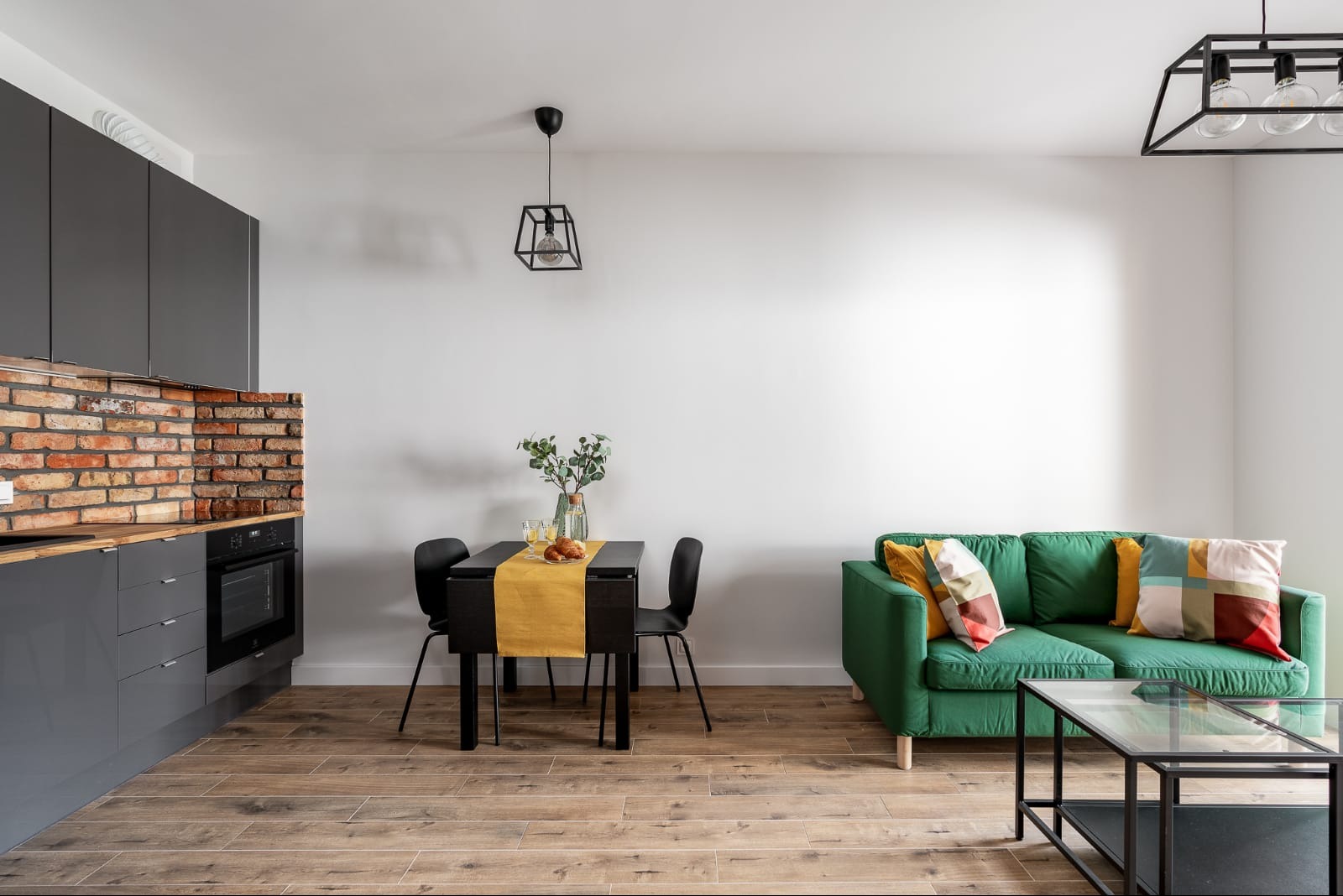 Jasne wnętrze mieszkania: szare szafki kuchenne z ceglanym backsplash'em, czarny stół z żółtym bieżnikiem, zielona sofa z kolorowymi poduszkami, drewnopodobna podłoga, czarne lampy sufitowe.