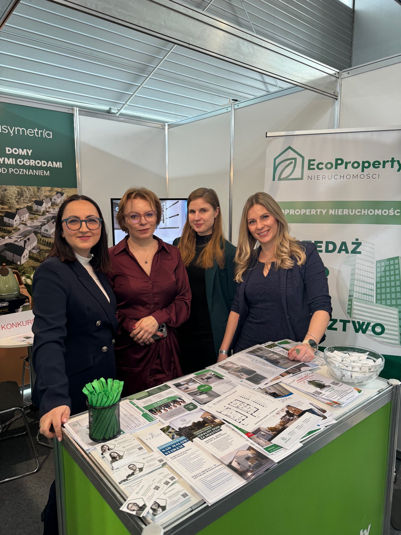 Stoisko EcoProperty na targach nieruchomości w Poznaniu: cztery kobiety w eleganckich strojach prezentują ulotki z ofertami domów i mieszkań na sprzedaż, na stole zielony pojemnik z długopisami...