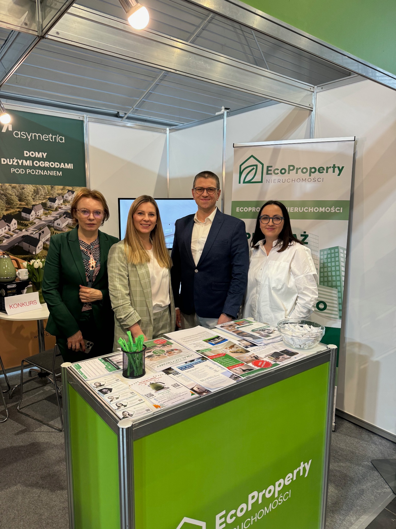 Stoisko targowe EcoProperty Nieruchomości z czterema osobami, ulotkami i banerem reklamowym na tle stoisk innych firm.