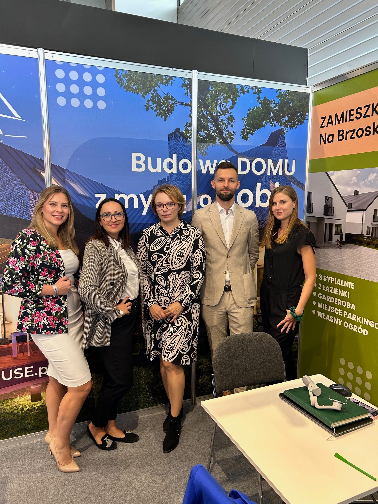 Zespół pracowników firmy z Poznania prezentuje się na tle stoiska targowego z wizualizacjami domów i hasłem 'Budowa w DOMU z my ob' oraz informacjami o ofercie 'Zamieszkaj na Brzosk'.