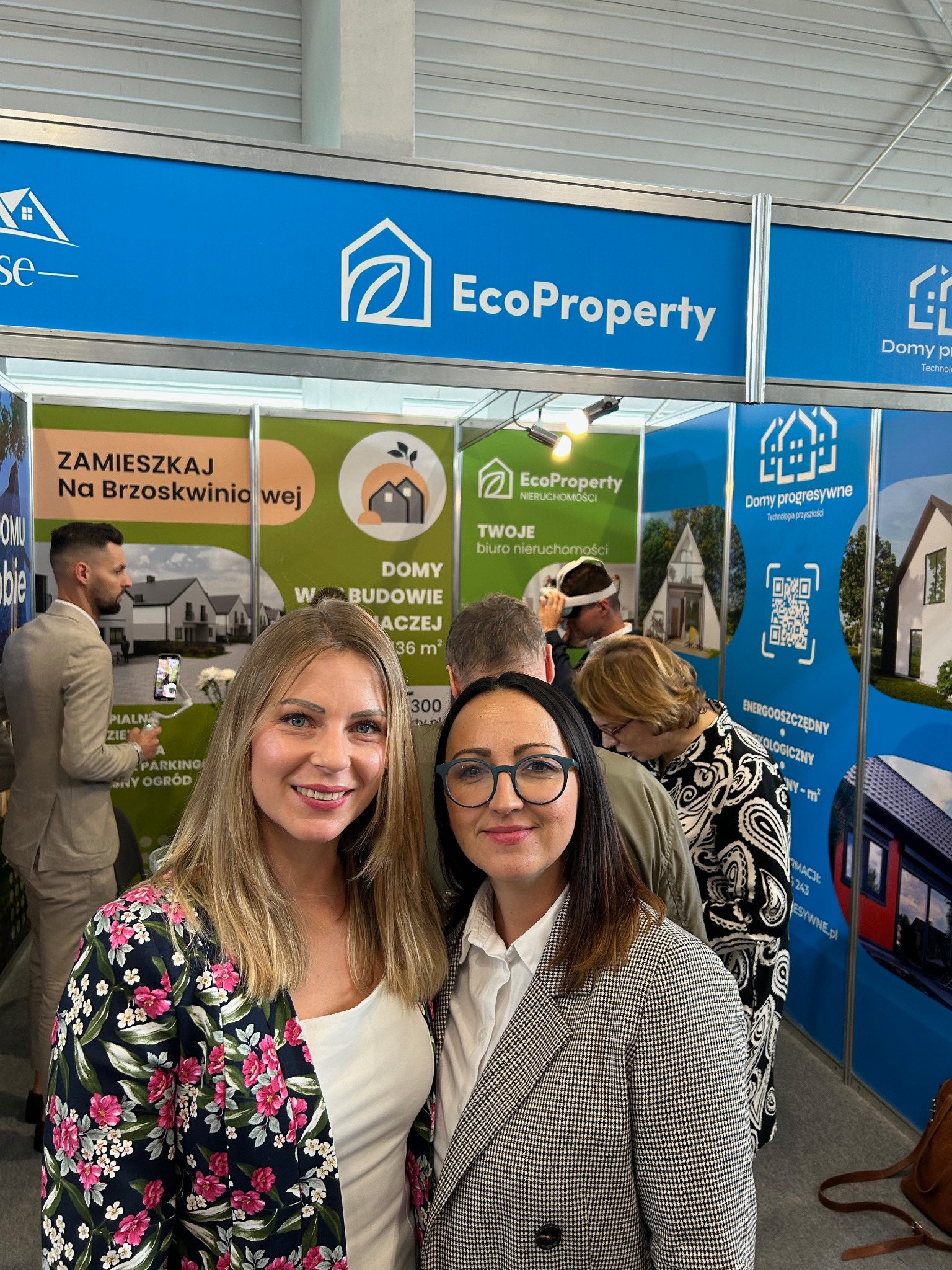 Dwie uśmiechnięte kobiety na tle stoiska targowego EcoProperty z wizualizacjami domów i informacjami o metrażu, z widocznym logo firmy i QR kodem.