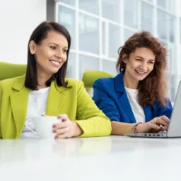 Dwie uśmiechnięte kobiety w biurze: brunetka w limonkowej marynarce trzyma biały kubek, a ruda w niebieskiej marynarce pracuje na laptopie Dell.