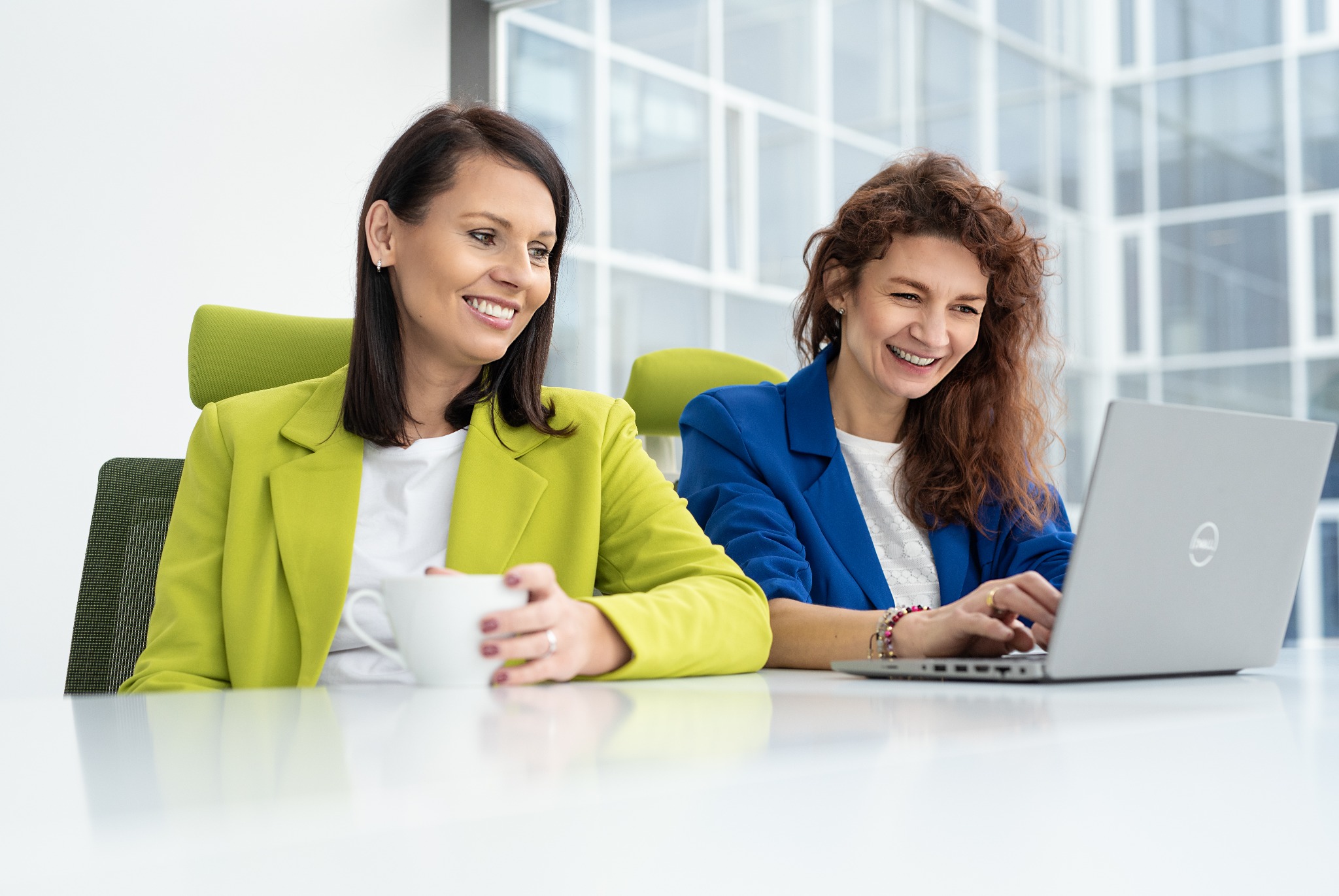 Dwie uśmiechnięte kobiety w biurze: brunetka w limonkowej marynarce trzyma biały kubek, a ruda w niebieskiej marynarce pracuje na laptopie Dell.
