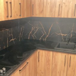 GrimzSa Remonty i wykończenia mieszkań - Kuchenna zabudowa z drewnopodobnymi szafkami i czarnym blatem oraz panelem ściennym z efektem marmuru z złotymi żyłami, widoczny czarny zlew i płyta indukcyjna.