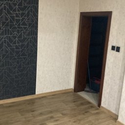 GrimzSa Remonty i wykończenia mieszkań - Pusty pokój po remoncie z panelami podłogowymi, wzorzystą tapetą na jednej ścianie i jasną tapetą na pozostałych ścianach, widoczne gniazdka elektryczne i lampa sufitowa.