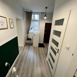 GrimzSa Remonty i wykończenia mieszkań - Korytarz po remoncie z szarą podłogą imitującą drewno, białe ściany z ciemnozielonym pasem, białe drzwi z przeszkleniami, widoczne dwa obrazy na ścianie i lampy sufitowe