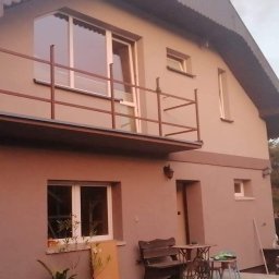 GrimzSa Remonty i wykończenia mieszkań - Dwupiętrowy dom po remoncie z balkonem, pomalowany na beżowo z brązowymi akcentami, widoczne okna i drzwi, na pierwszym planie roślinność i meble ogrodowe.