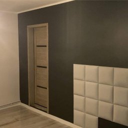 GrimzSa Remonty i wykończenia mieszkań - Pokój po remoncie z szarą ścianą, drewnianymi drzwiami z czarnymi paskami, panelami tapicerowanymi na ścianie i grzejnikiem pod oknem.