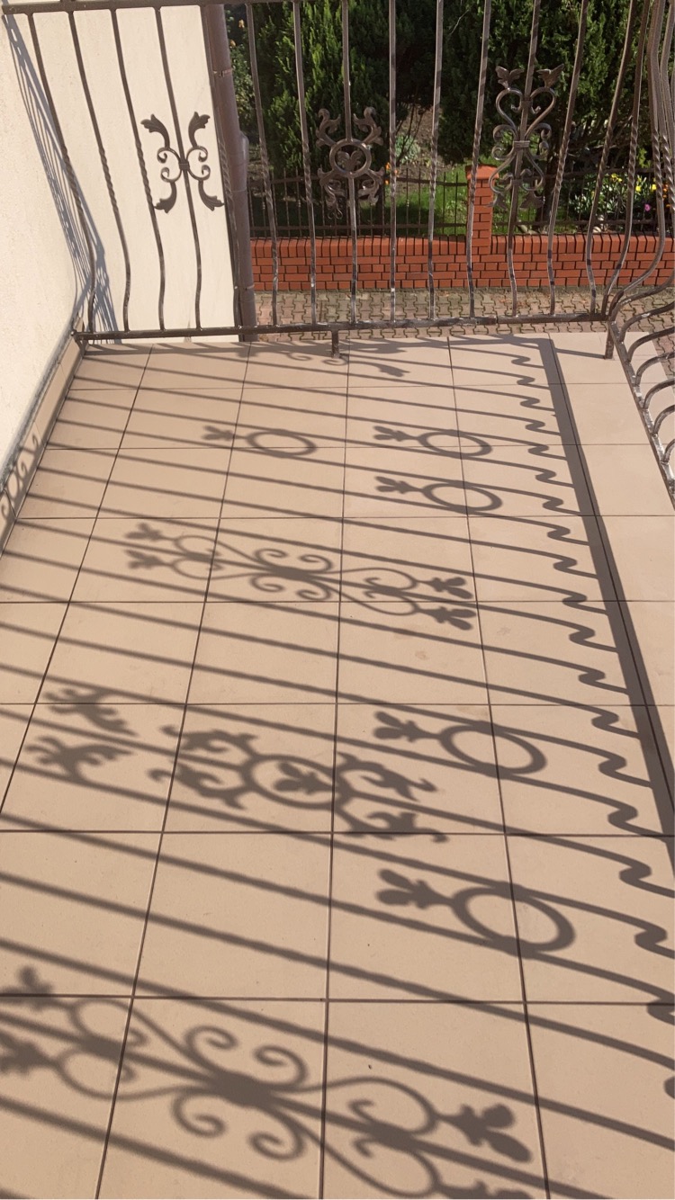 Kafelki na balkonie w ciepłym odcieniu, oświetlone promieniami słońca rzucającymi ażurowe cienie metalowej balustrady.