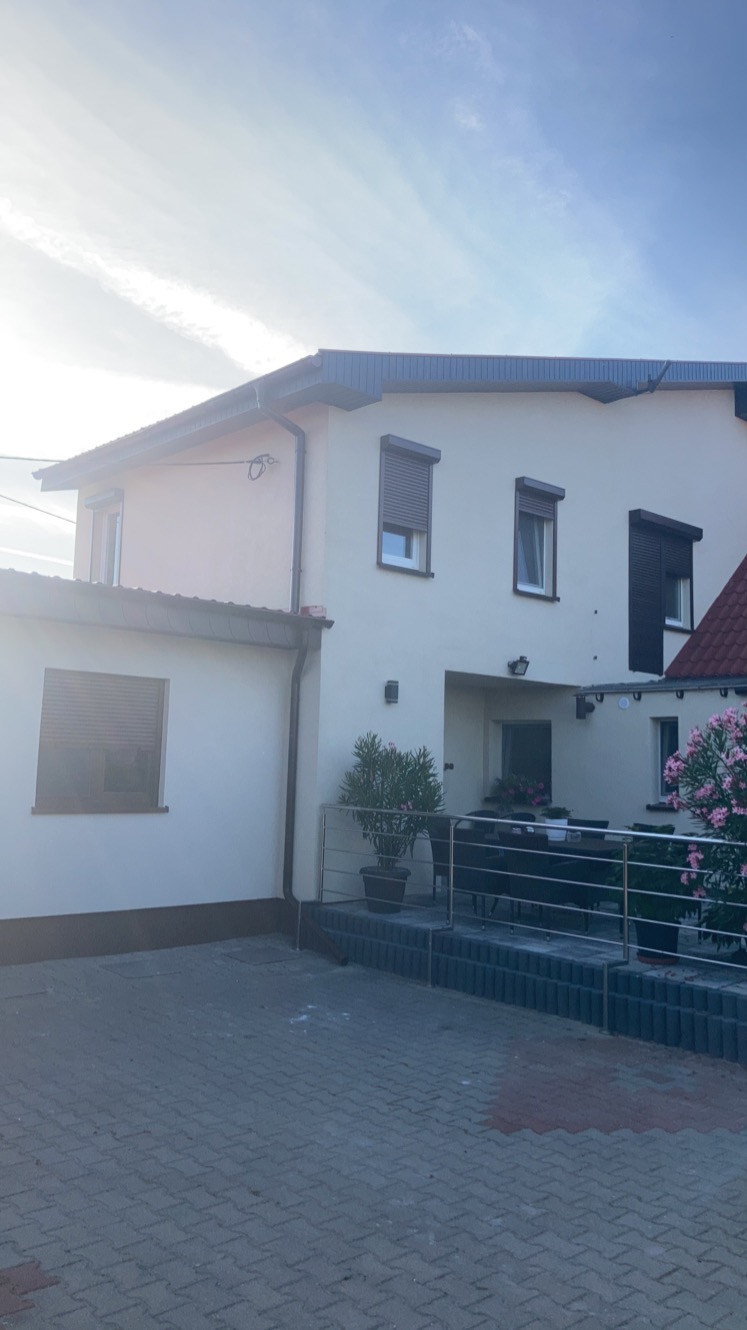 Jasna elewacja domu jednorodzinnego z brązowymi roletami zewnętrznymi, widok z poziomu kostki brukowej przed domem.