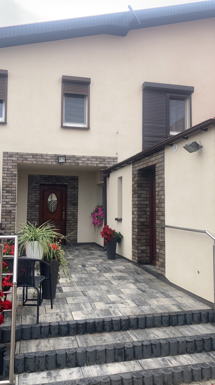 Elewacja domu jednorodzinnego po renowacji z nową kostką brukową na podjeździe, widoczne okna z roletami zewnętrznymi i dekoracyjne donice z kwiatami.