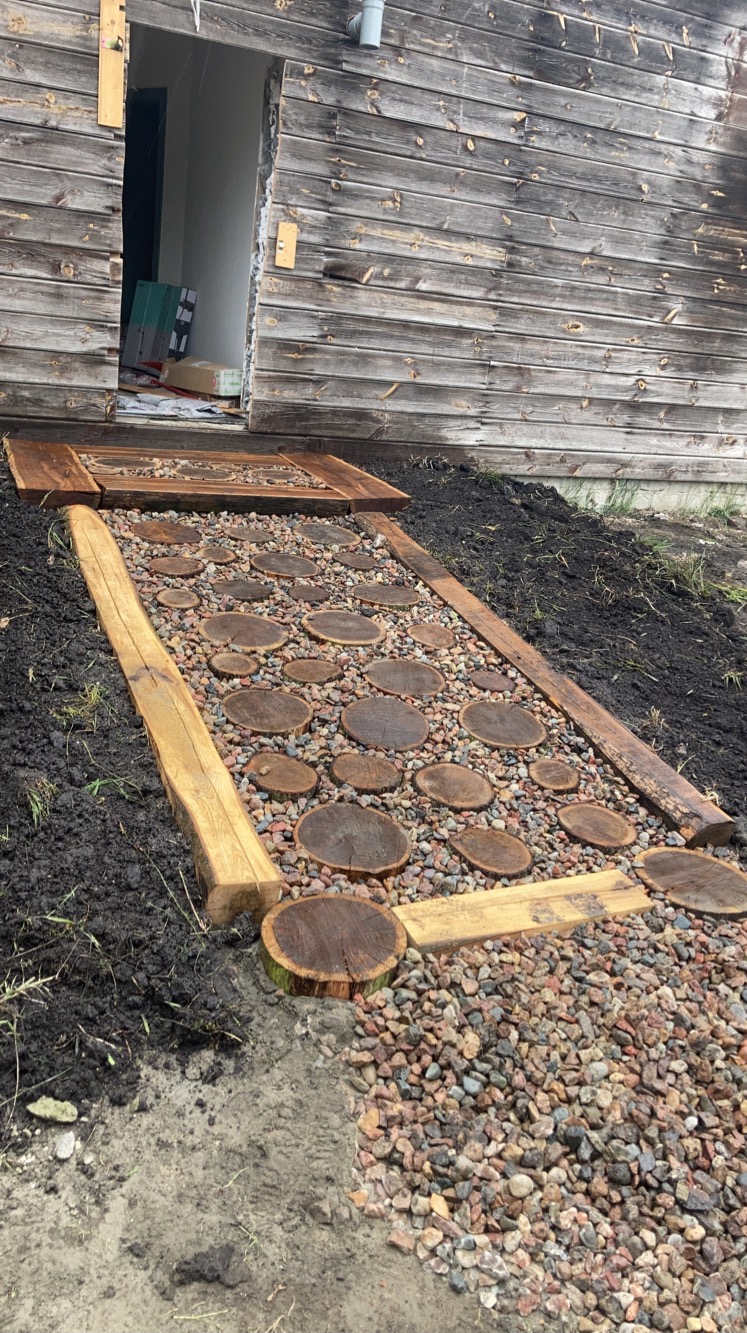 Ścieżka w ogrodzie wyłożona okrągłymi plastrami drewna i drobnymi kamieniami, obramowana drewnianymi belkami, prowadząca do wejścia do drewnianego domu.