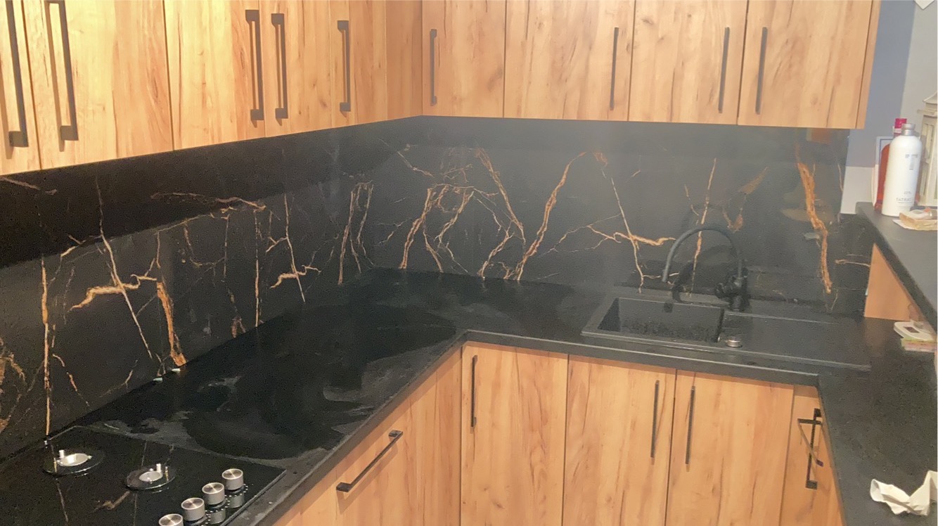 Kuchenna zabudowa z drewnopodobnymi szafkami i czarnym blatem oraz panelem ściennym z efektem marmuru z złotymi żyłami, widoczny czarny zlew i płyta indukcyjna.