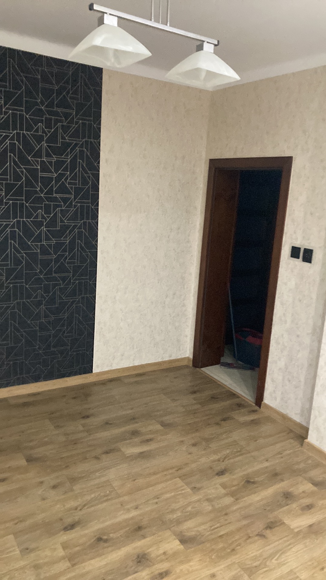 Pusty pokój po remoncie z panelami podłogowymi, wzorzystą tapetą na jednej ścianie i jasną tapetą na pozostałych ścianach, widoczne gniazdka elektryczne i lampa sufitowa.