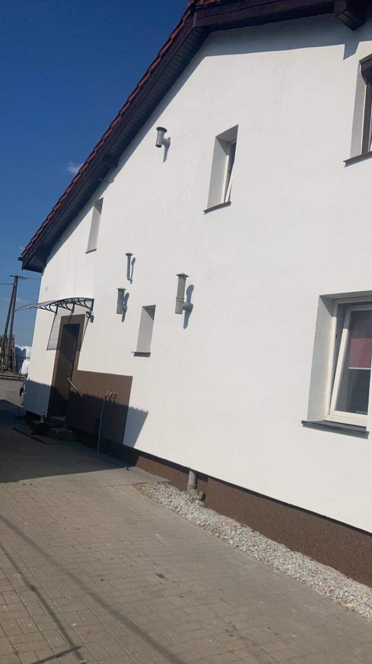 Boczny widok odnowionego domu z białą elewacją, ciemnobrązowym dachem i cokolem, widoczne okna i rury wentylacyjne.