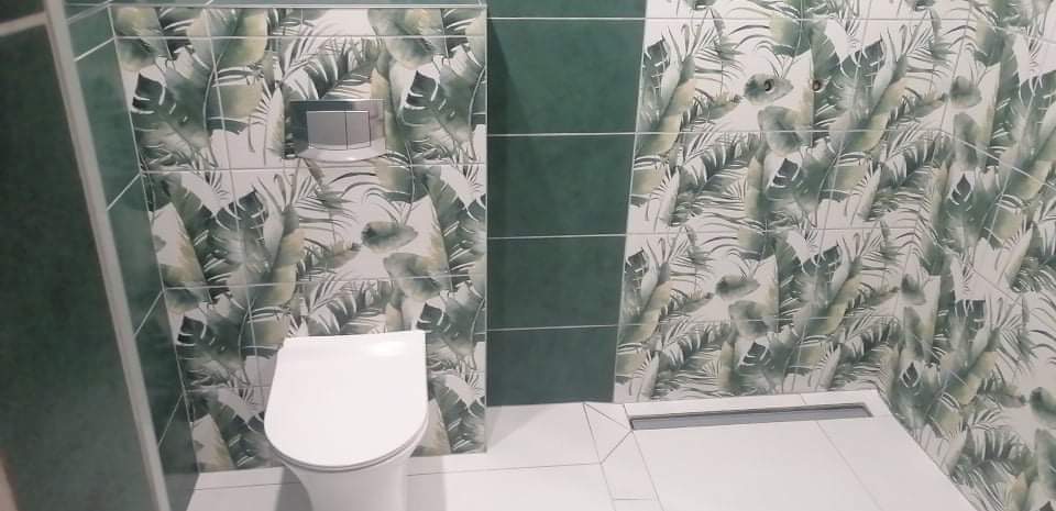 Nowoczesna toaleta z podwieszaną muszlą, obudowana płytkami w motywie tropikalnych liści, z akcentami w postaci pionowych pasów z jednolitych, ciemnozielonych płytek.