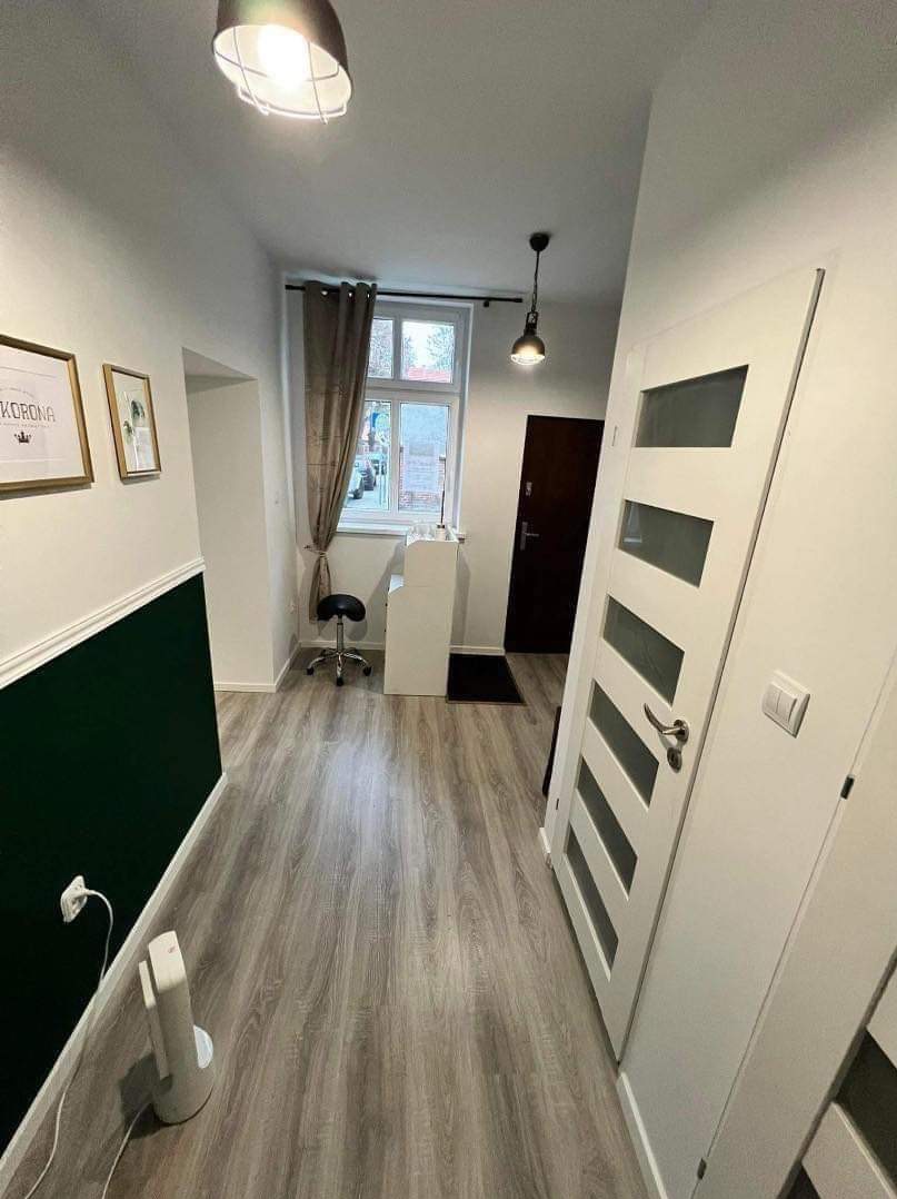 Korytarz po remoncie z szarą podłogą imitującą drewno, białe ściany z ciemnozielonym pasem, białe drzwi z przeszkleniami, widoczne dwa obrazy na ścianie i lampy sufitowe