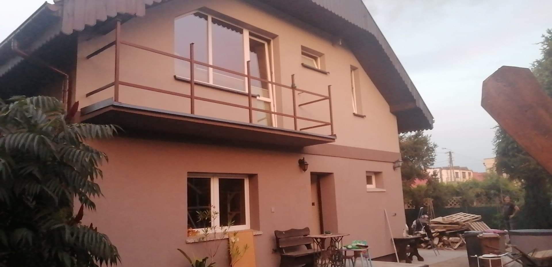 Dwupiętrowy dom po remoncie z balkonem, pomalowany na beżowo z brązowymi akcentami, widoczne okna i drzwi, na pierwszym planie roślinność i meble ogrodowe.