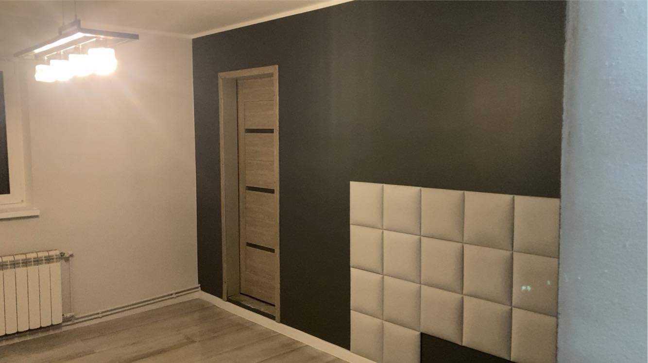 Pokój po remoncie z szarą ścianą, drewnianymi drzwiami z czarnymi paskami, panelami tapicerowanymi na ścianie i grzejnikiem pod oknem.