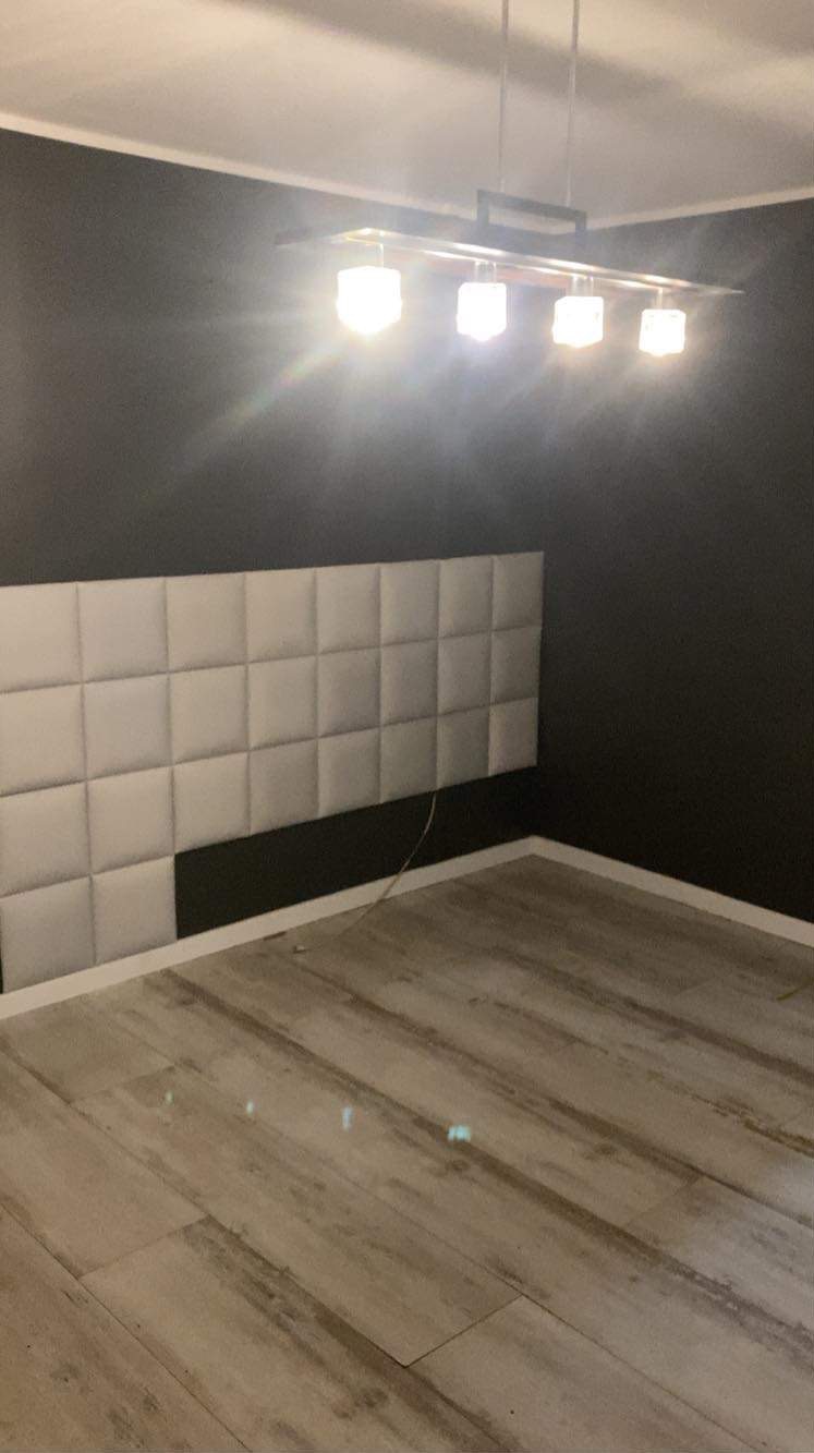 Pusta, ciemnoszara ściana z zamontowanym panelem tapicerowanym w formie jasnoszarych kwadratów, podświetlona przez nowoczesną lampę sufitową z czterema punktami świetlnymi, na podłodze panele...