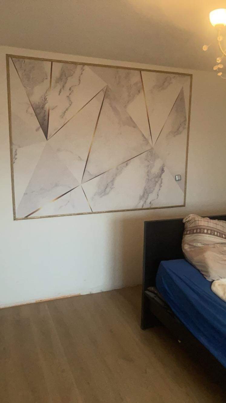 Geometryczny wzór marmuru z złotymi liniami na ścianie w sypialni, widoczny fragment łóżka z niebieską pościelą i poduszką.